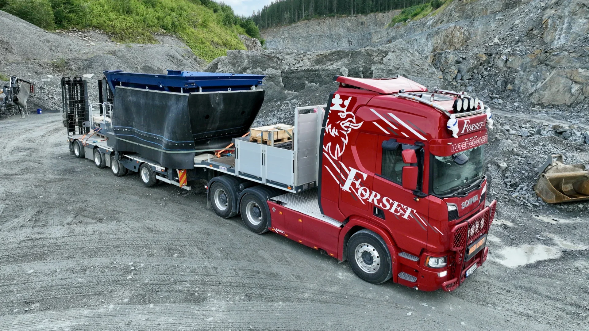 Red Forset transport truck hauling a large black industrial hull on a multi-axle trailer in a rocky quarry setting.